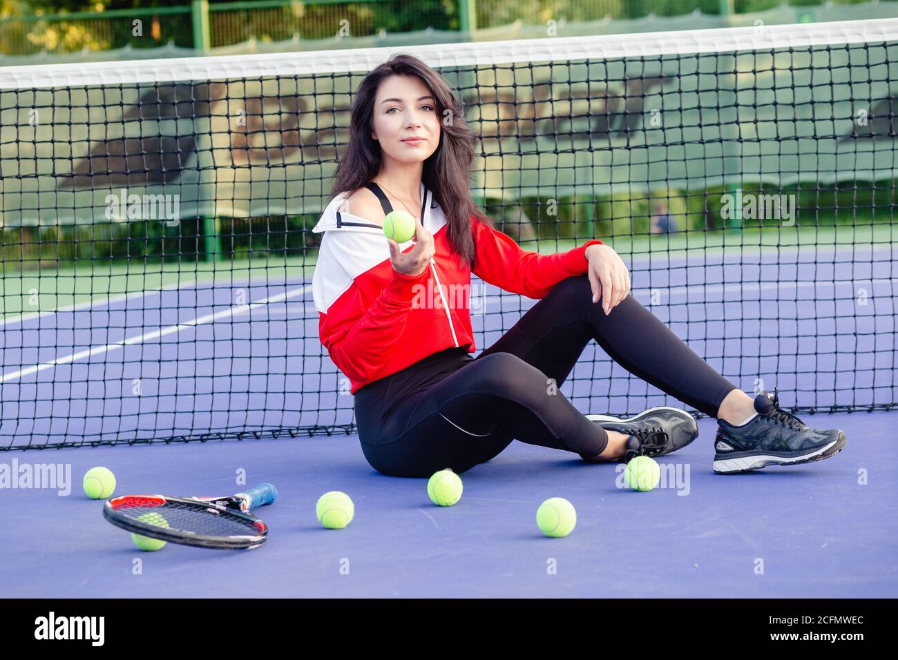 Giovane bella donna seduta appoggiata sulla rete da tennis, riposandosi rilassandosi dopo l'allenamento. Ritratto di sportswoman sul campo da tennis blu. Vita sana. Foto Stock