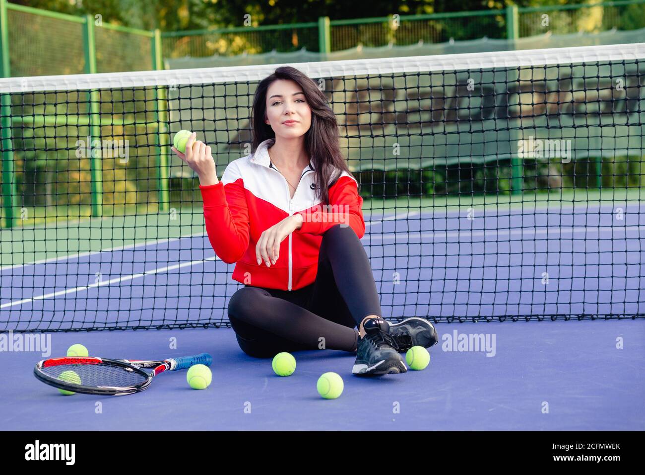 Giovane bella donna seduta appoggiata sulla rete da tennis, riposandosi rilassandosi dopo l'allenamento. Ritratto di sportswoman sul campo da tennis blu. Vita sana. Foto Stock