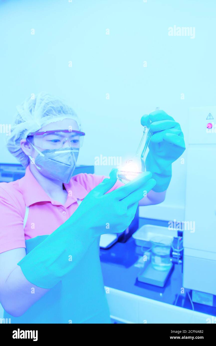Una donna asiatica che indossa una maschera chirurgica che tiene un matraccio volumetrico o una provetta che fa la ricerca in un laboratorio. Messa a fuoco morbida su fl. Volumetrica Foto Stock