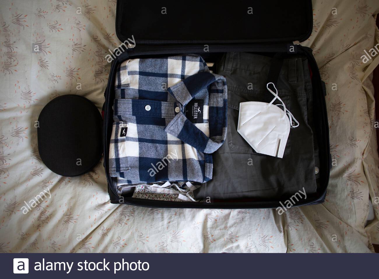 La borsa da viaggio di una persona si apre e imballata su un letto prima della partenza con una camicia e una maschera bianca anti-Covid19 prominente nella foto. Foto Stock
