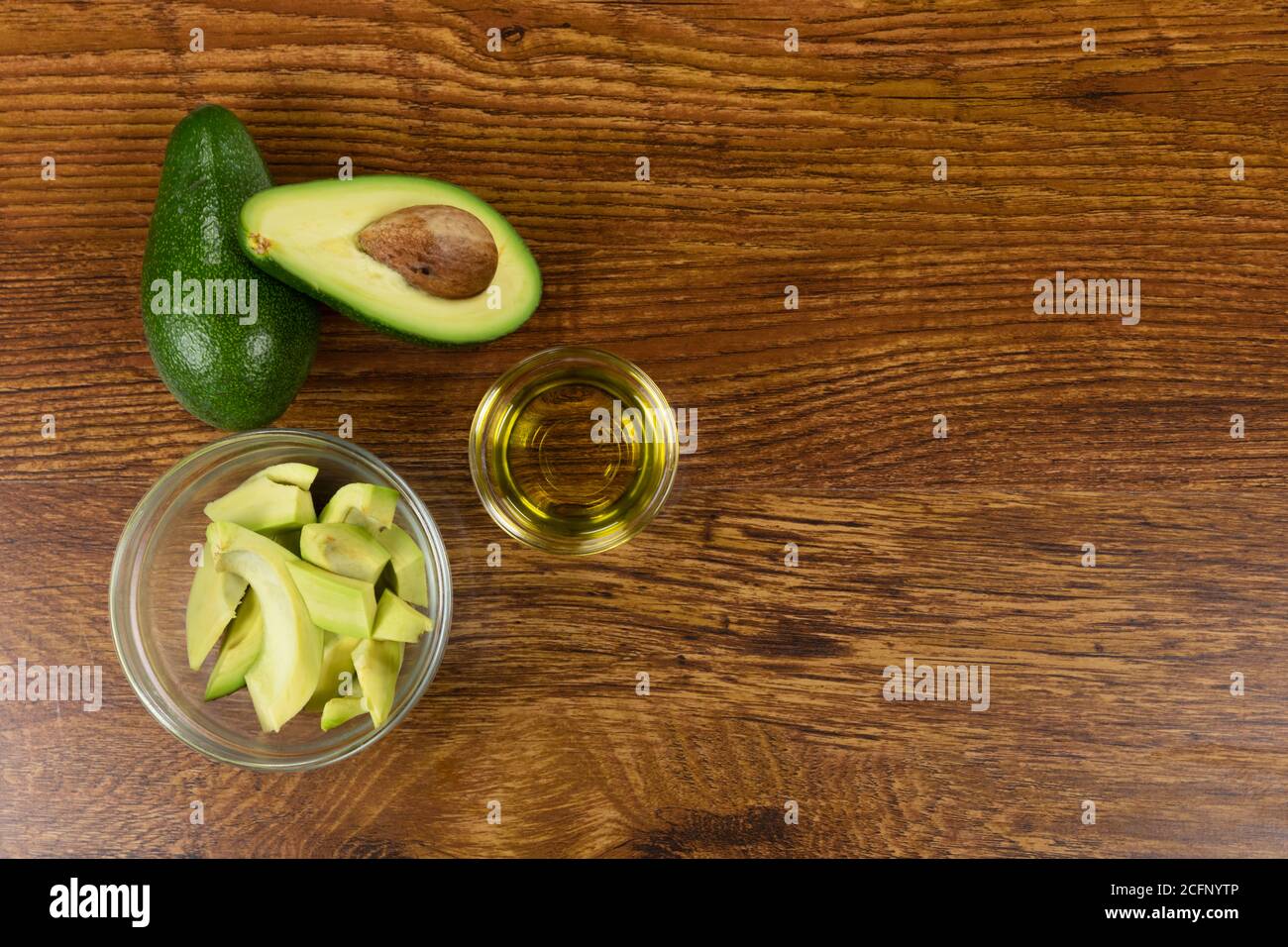 Vista di due avocado, bottiglia di olio d'oliva e avocado tagliato in una ciotola su sfondo tavola di legno Foto Stock