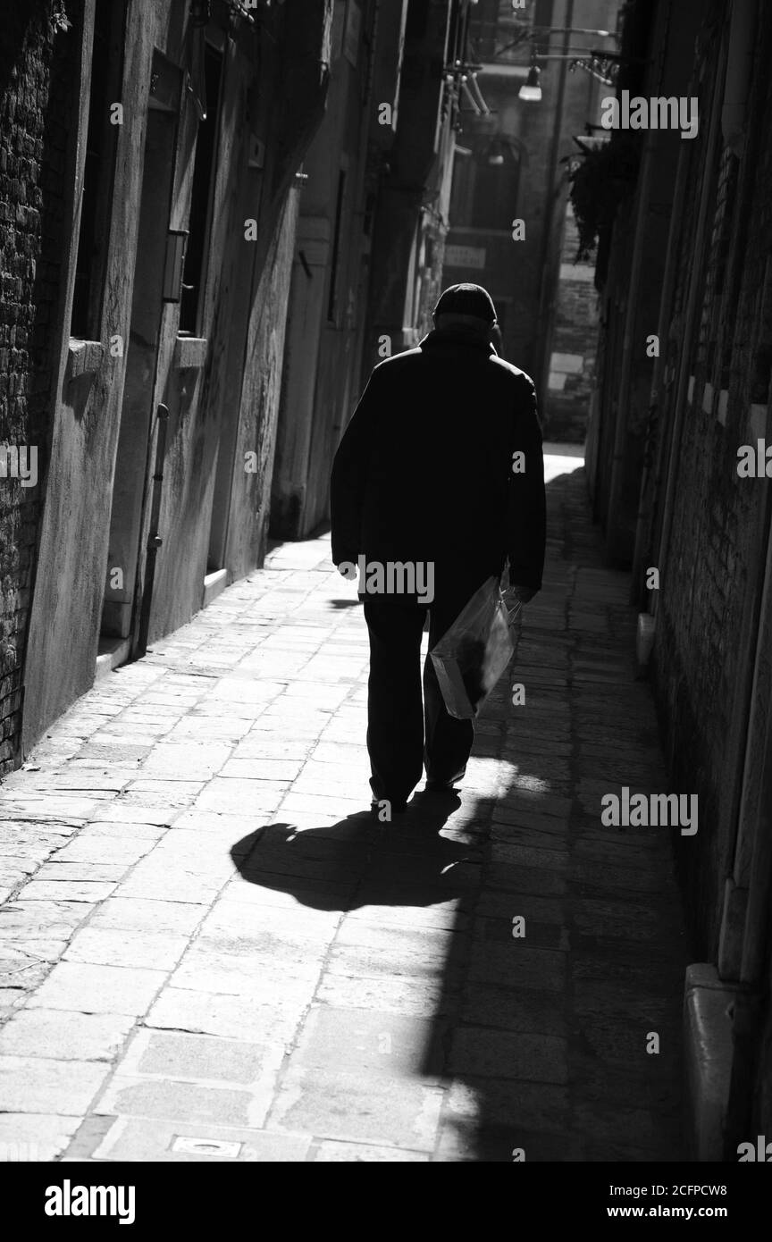Silhouette di vecchio uomo che va su strada stretta in raggi di sole splendenti. (Venezia, Italia) un gioco di luce e ombra. Punti luce. Foto in bianco e nero. Foto Stock