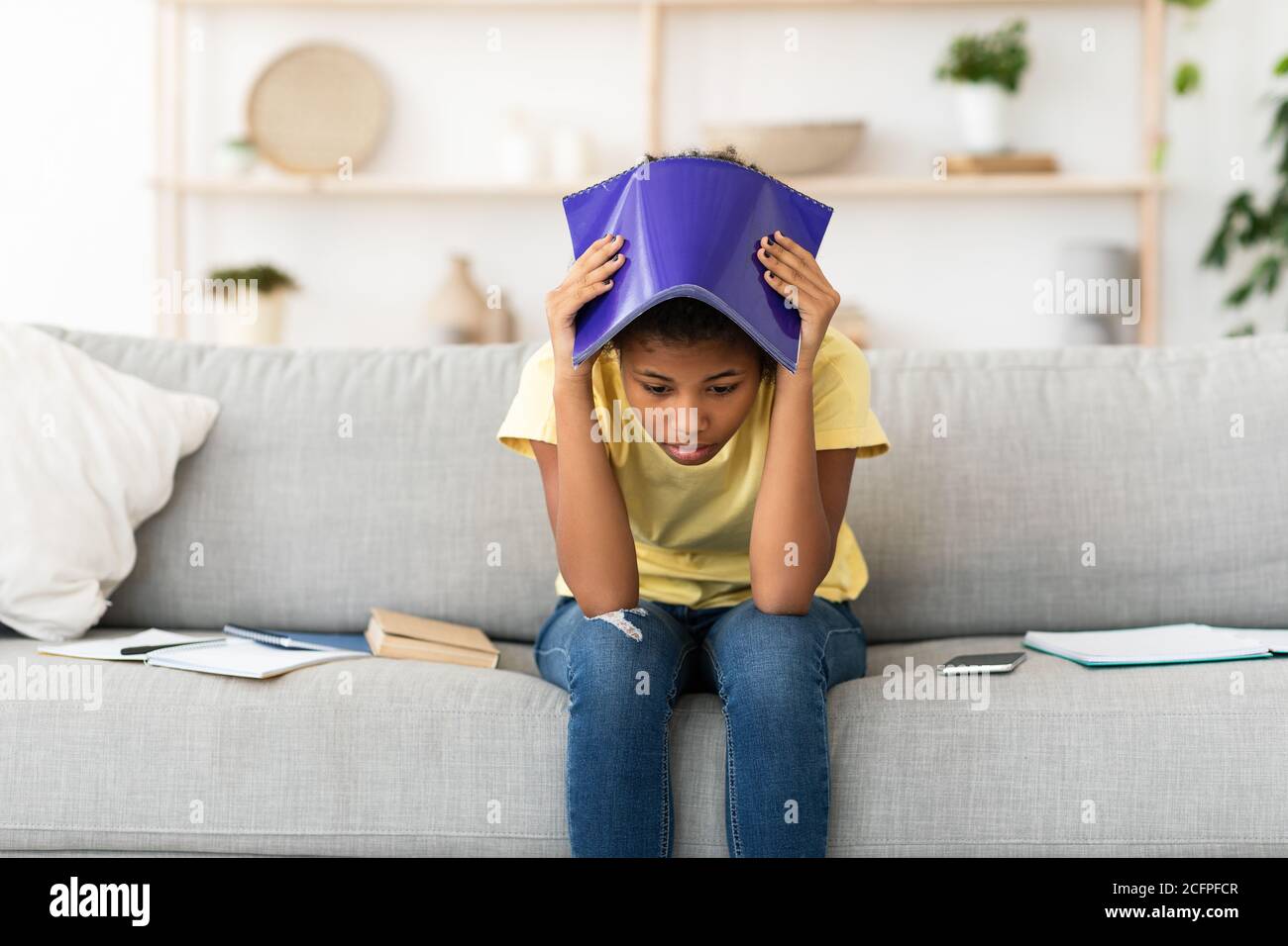 Ragazza sconvolto coprendo testa con esercizio-Libro seduta su divano interno Foto Stock
