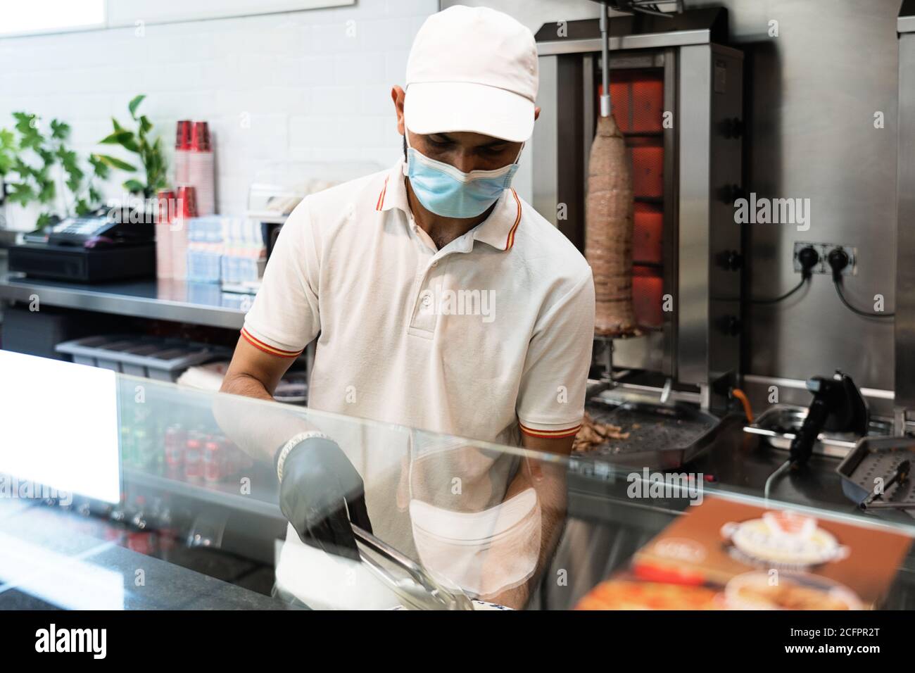 Chef in uniforme, tagliando e preparando kebab di pollo e carne donatrice utilizzando una maschera di prevenzione viso presso un fast food stand Foto Stock