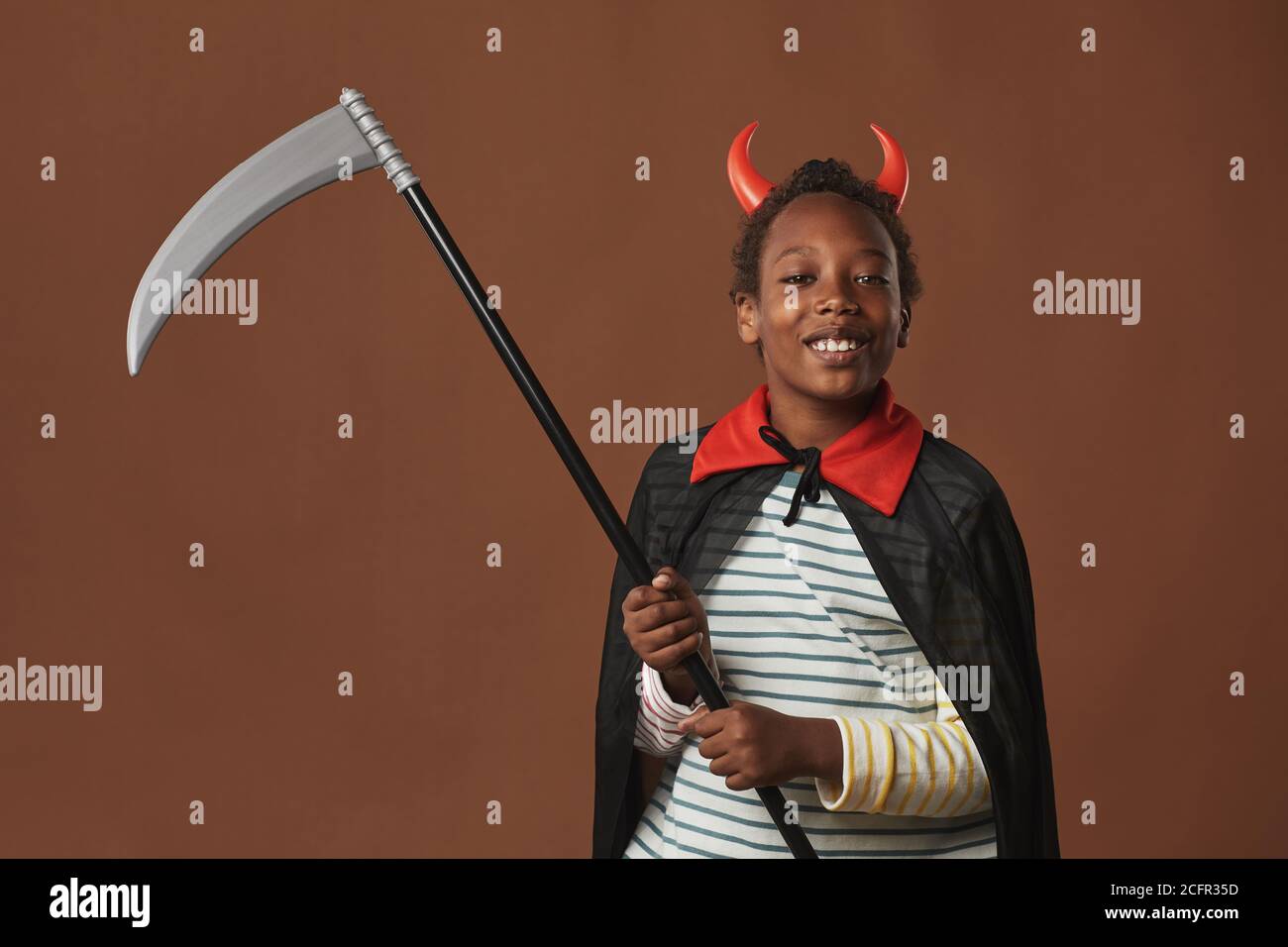 Studio ritratto di allegro ragazzo preteen afroamericano con corna rosse sulla testa e mantello in costume che tiene la falce guardando la macchina fotografica sorridente, sfondo marrone Foto Stock