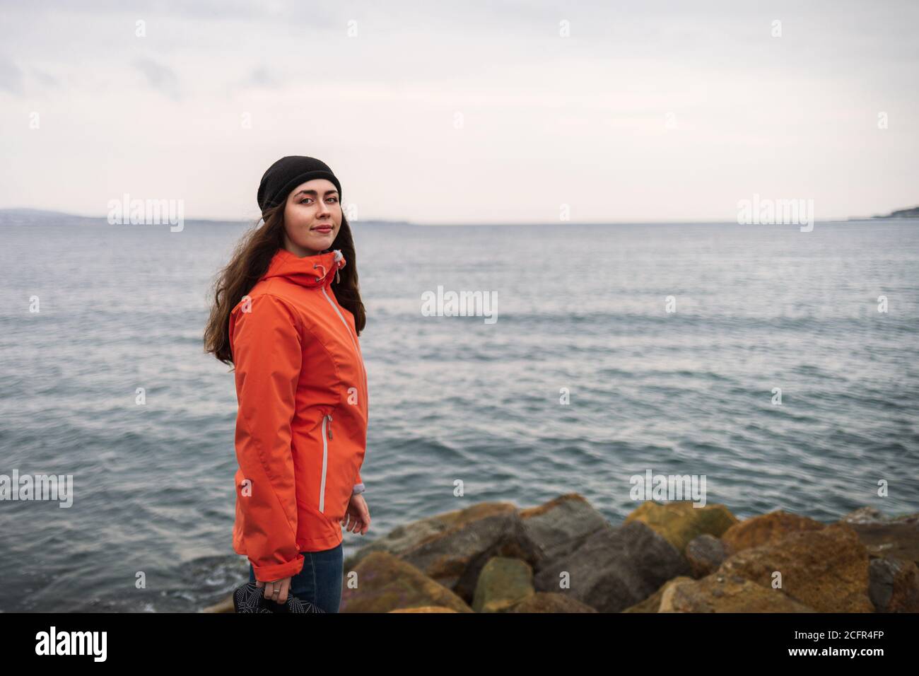 Donna brunetta con capelli lunghi in un cappello e una giacca arancione, in posa sulla spiaggia. Sullo sfondo il mare e la linea dell'orizzonte. Copiare spazio e chiudere Foto Stock