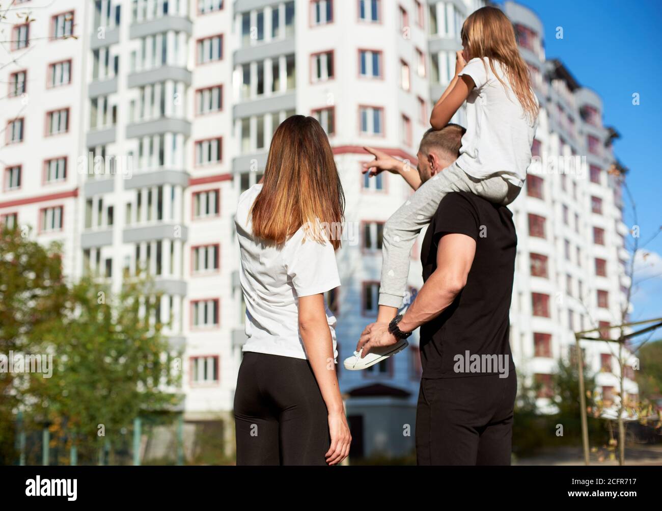 La famiglia sportiva di tre persone sta in piedi con la schiena alla fotocamera e guardando un grande blocco di appartamenti, la ragazza è seduta sulle spalle del padre. L'uomo sta puntando su un edificio con un grande blocco di appartamenti Foto Stock