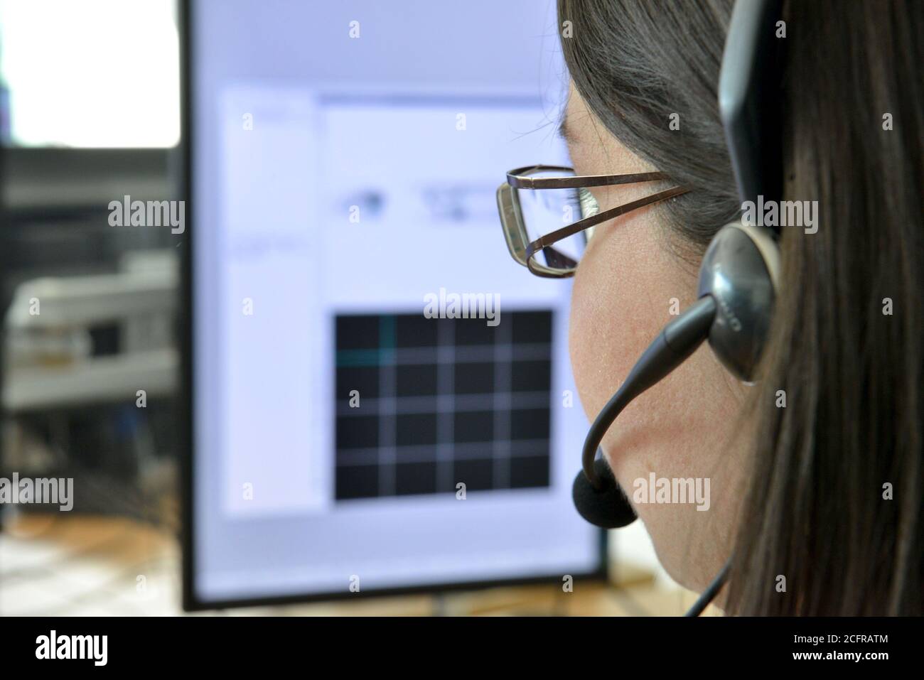 Ritratto stretto di una giovane donna con occhiali, vista da dietro, con un auricolare e microfono davanti allo schermo del suo computer Foto Stock