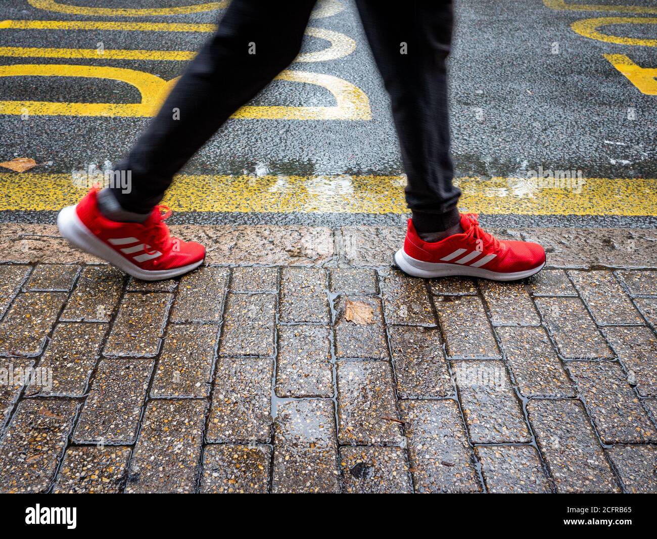 Persone gambe e piedi che camminano dal marciapiede su un giornata piovosa bagnata con scarpe da ginnastica a righe rosse Foto Stock