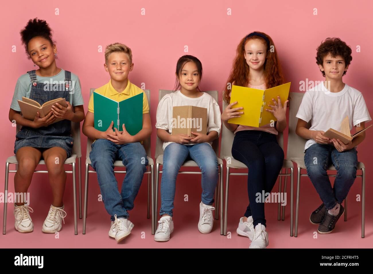 Ritratto a lunghezza intera degli scolari con materiale educativo seduto sopra sedie su sfondo rosa Foto Stock