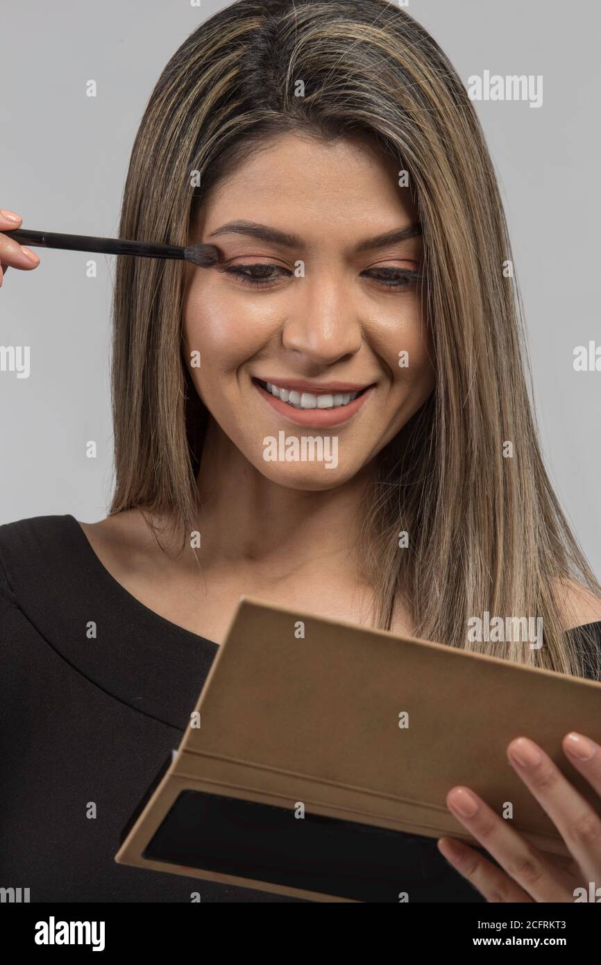 faccia di donna latina con biondo e capelli lunghi che mettono sul trucco con l'altra mano che tiene uno specchio, studio Foto Stock