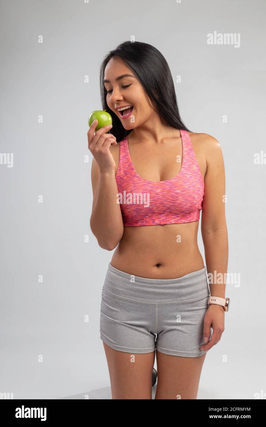 donna giovane, snella e latina che mangia una mela verde, usando il top rosa ang corto grigio, in studio con sfondo grigio Foto Stock