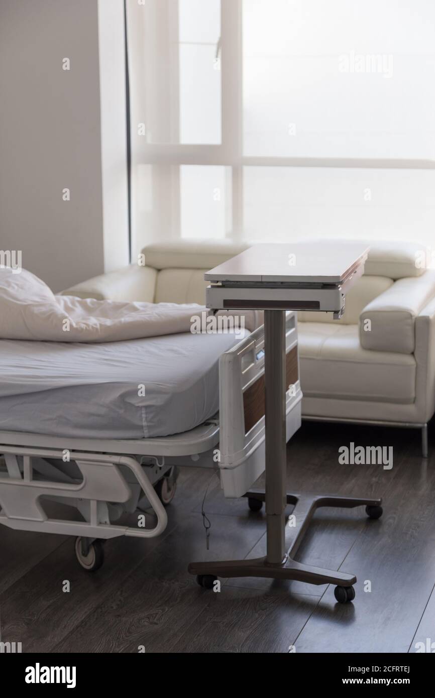 gurney dell'ospedale in camera con lenzuola bianche, un divano e un tavolo mobile accanto a una finestra Foto Stock