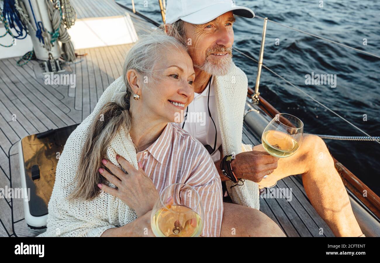 Coppia senior avvolta con plaid e seduta su un ponte di yacht con bocali di vino. Due persone affettuose e mature che si siedono insieme su una barca a vela e che guardano Foto Stock