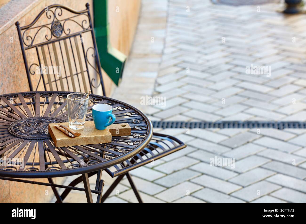 Street still life di un elegante tavolo in metallo con una tazza turchese, un bicchiere con un cucchiaio di metallo su un supporto di legno, sullo sfondo di pietre di pavimentazione i Foto Stock