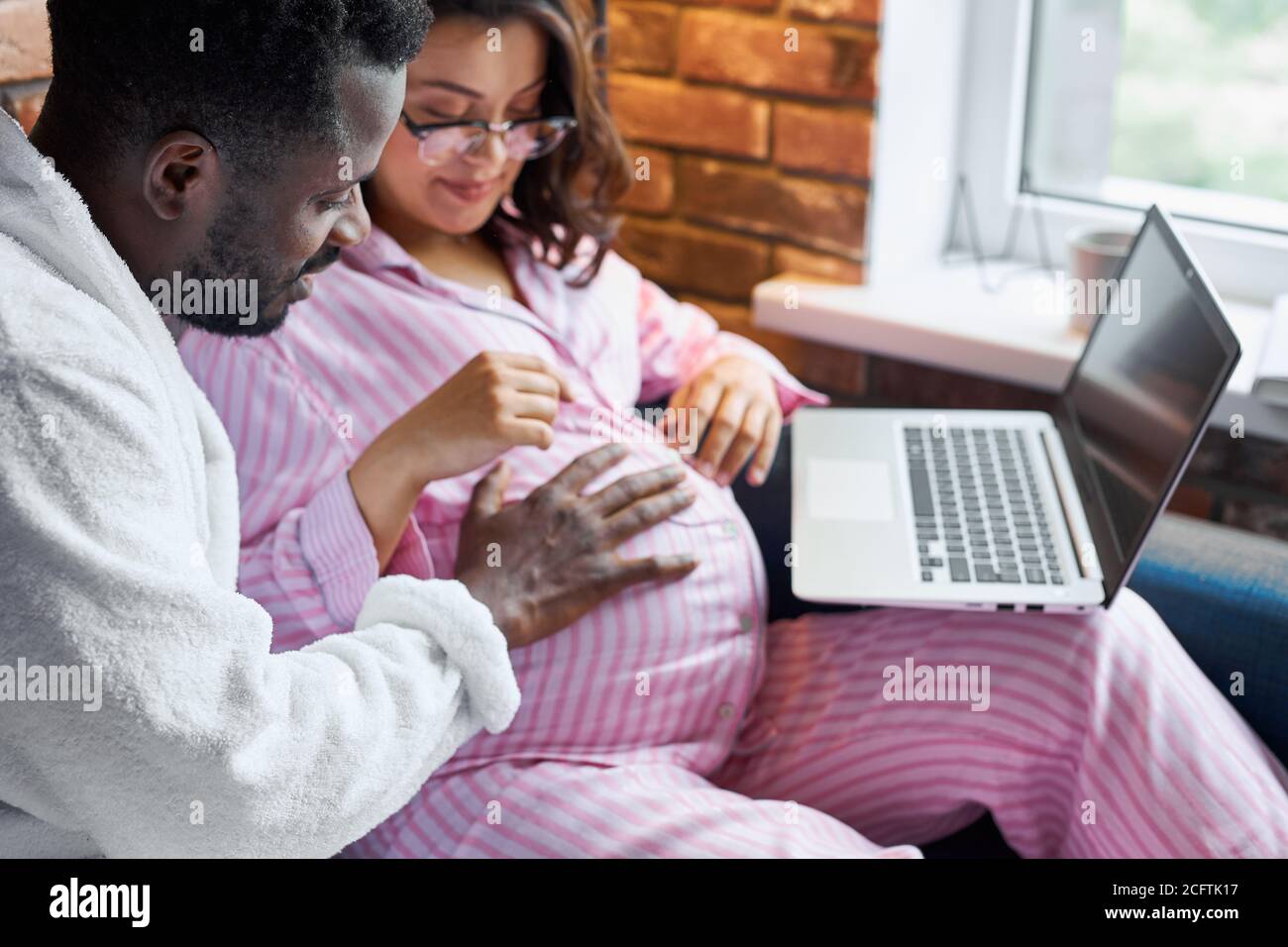 l'uomo nero è gentile con la sua moglie caucasica incinta, giovane maschio ictus pancia, donna in pigiama si siede con laptop a casa, innamorato Foto Stock