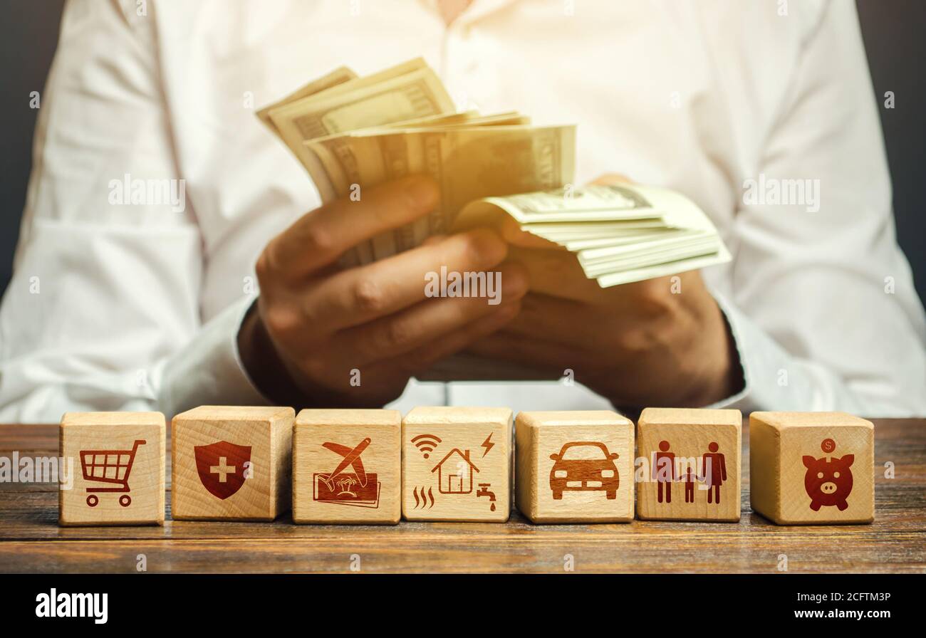 Un uomo conta soldi e blocchi con gli attributi dei bisogni della famiglia. Concetto di budget familiare, spese di bollette, vacanza e trasporto. Stipendio ottimale, Foto Stock