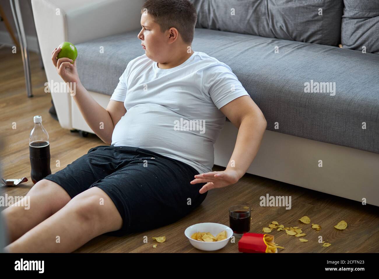 fat boy ha deciso di mangiare cibo sano, dire no al fast food. si siede sul pavimento in abbigliamento domestico, scegliere e mangiare. concetto di nutrizione Foto Stock