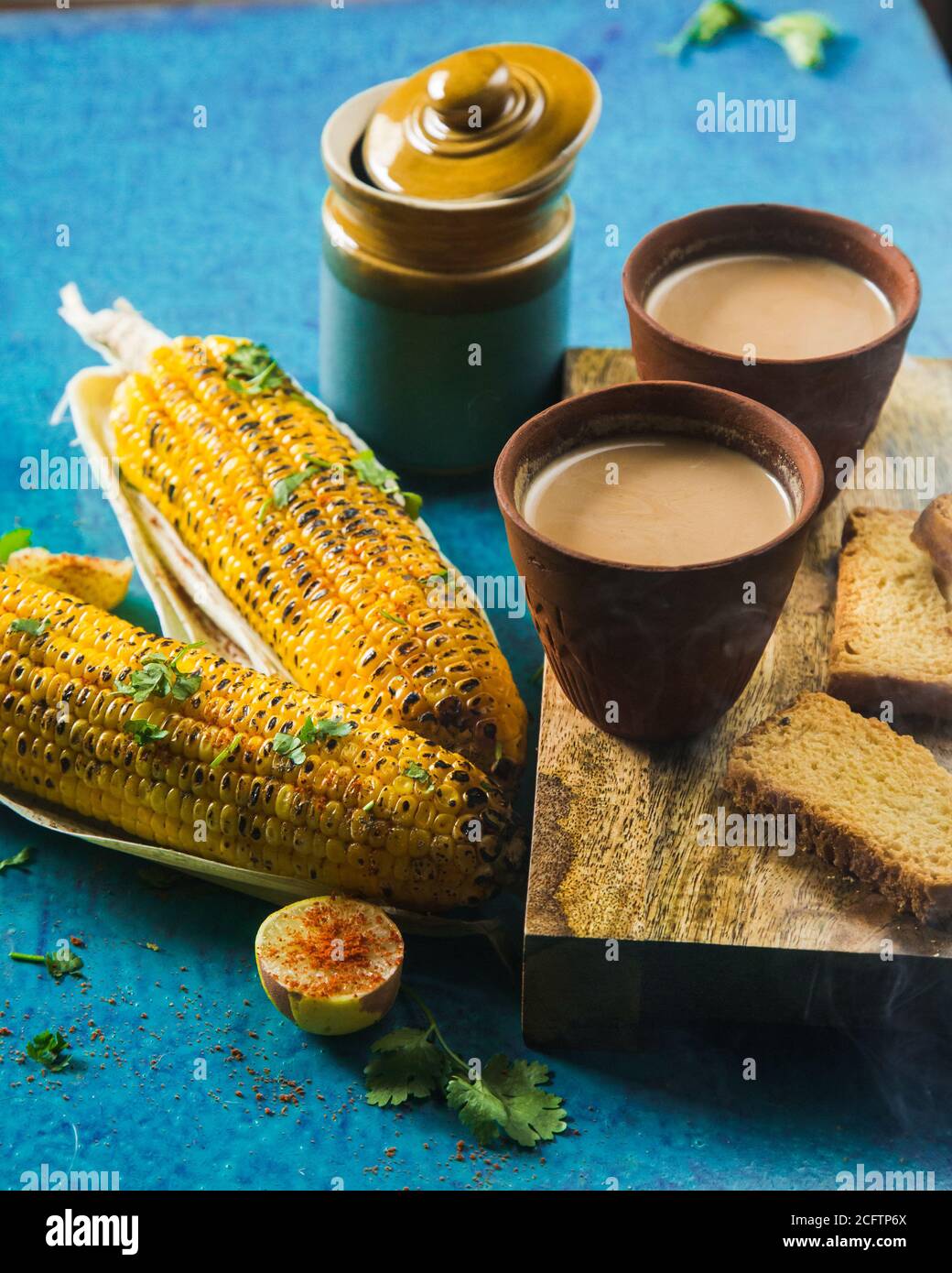 Vista dall'alto del tè al latte caldo preparato con granoturco arrosto alla griglia e pane tostato fresco. Deliziosi corncobs grigliati e due tazze di Masala Chai, un mon indiano ideale Foto Stock