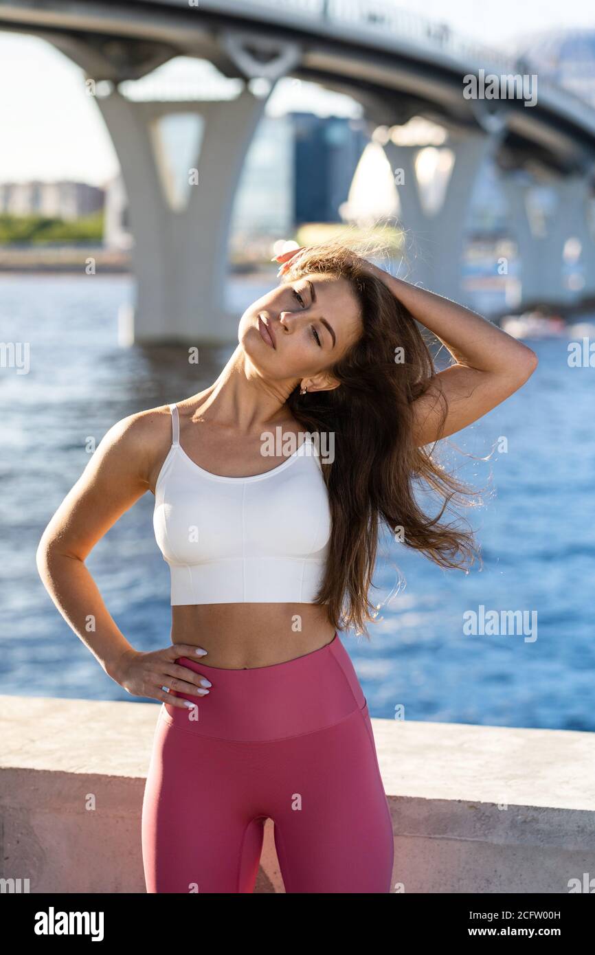 Adatta giovane donna atletica che allunga il corpo sull'argine. Fitness femminile in gambe rosa facendo allenamento caldo per i muscoli del collo all'aperto. Foto Stock