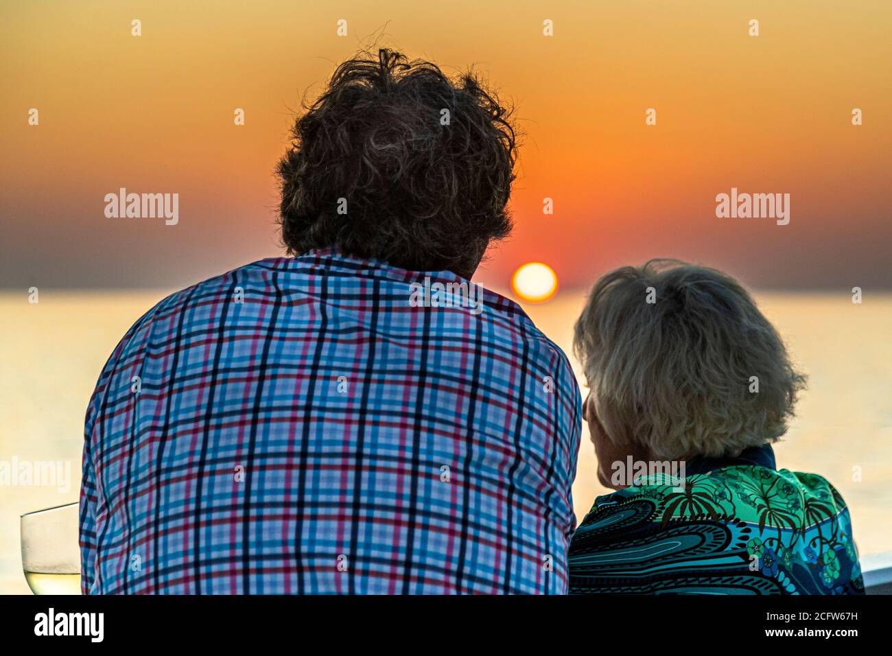 Una coppia seduta osserva il tramonto sul mare al largo delle isole della Sonda, Indonesia, con un bicchiere di vino bianco a portata di mano. Foto Stock
