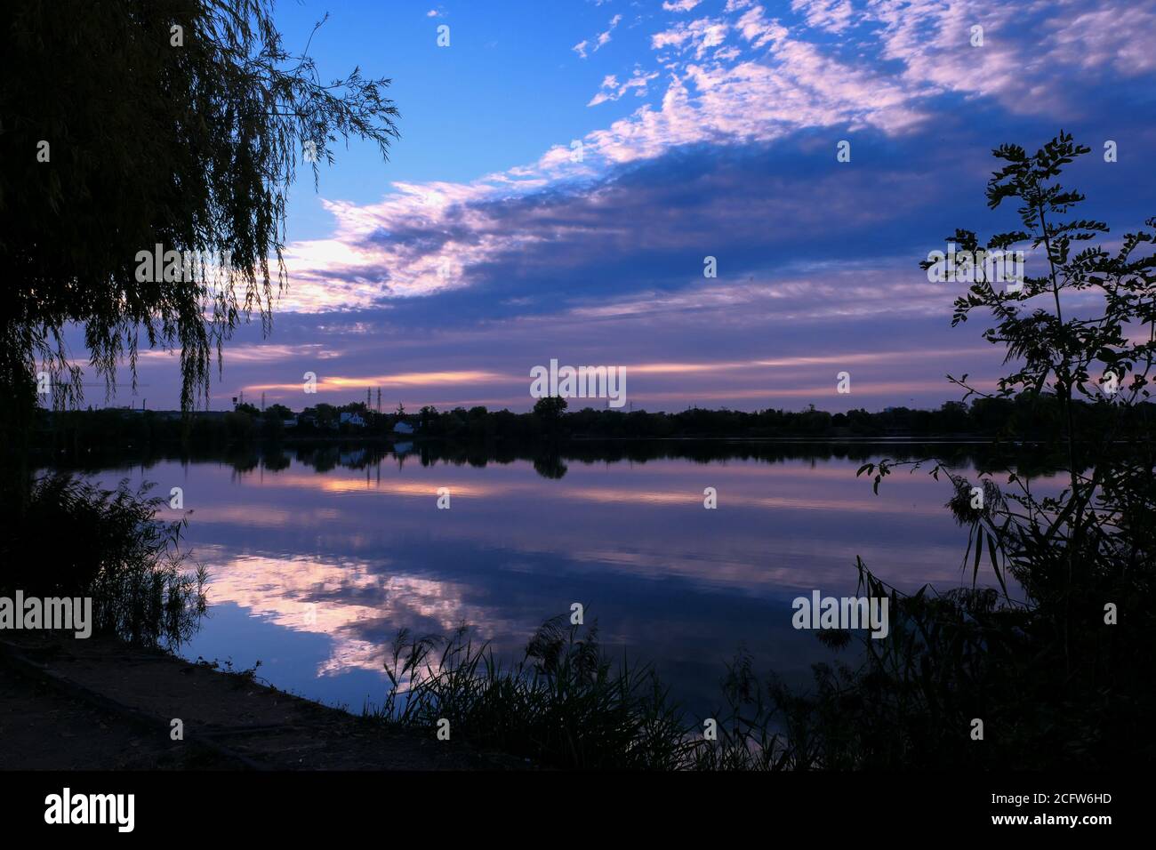 Simmetria del cielo in un lago all'alba. Nuvole che si riflettono sull'acqua. Paesaggio di vacanza al mare. Silhouette di vegetazioni in primo piano. Foto Stock