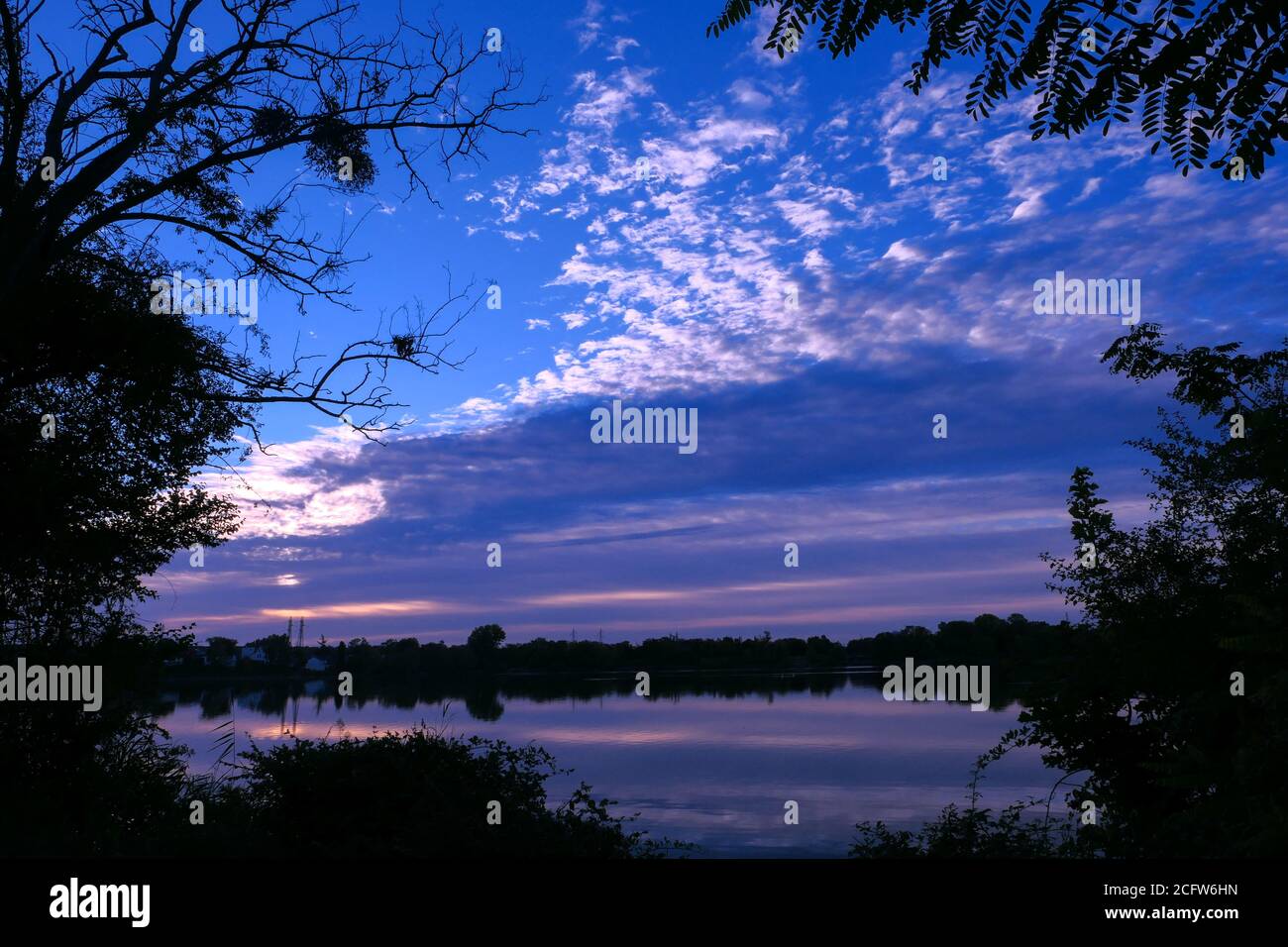 Simmetria del cielo in un lago all'alba. Nuvole che si riflettono sull'acqua. Paesaggio di vacanza al mare. Silhouette di vegetazioni in primo piano. Foto Stock
