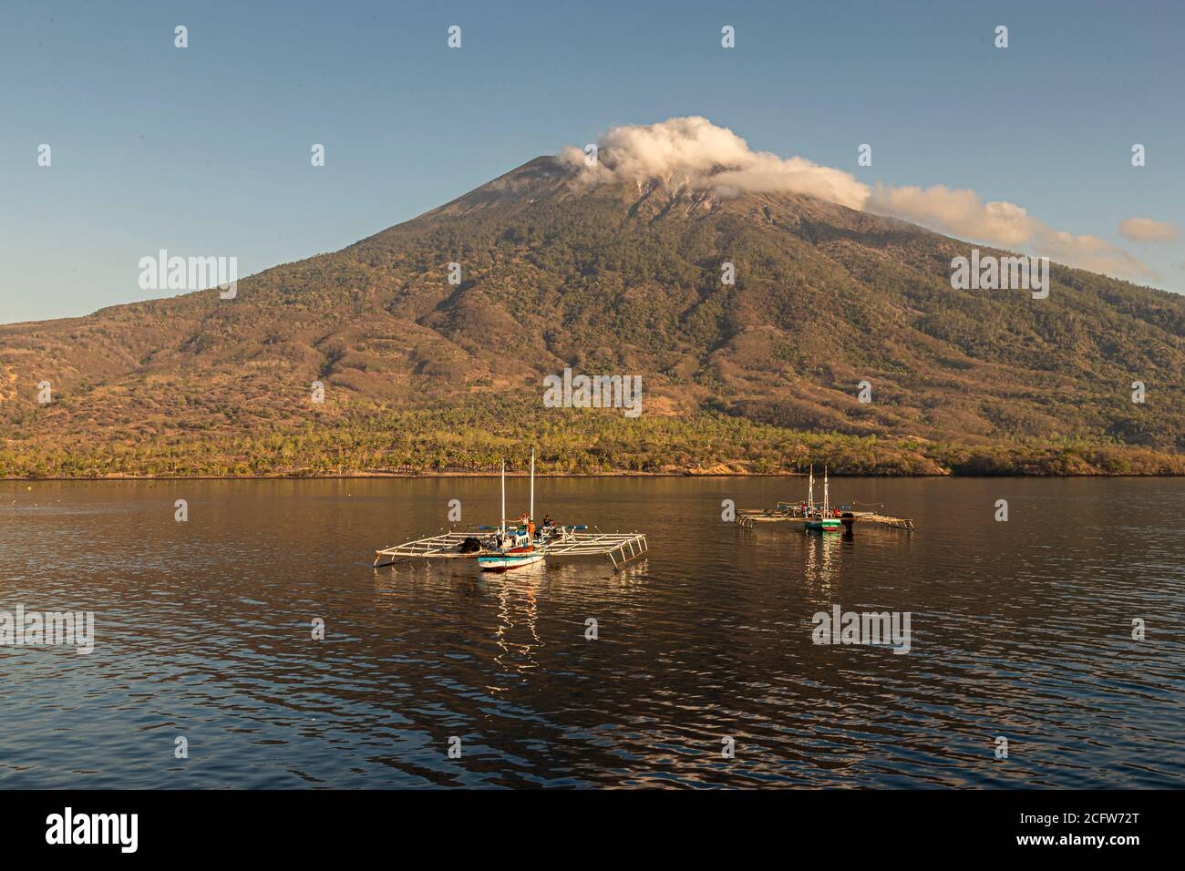 Il vulcano Ile Lewotolok si erge sopra due barche a bilanciere con telai a rete di sollevamento al largo della costa di Lembata, Indonesia, una nuvola avvolge la sua cima. Foto Stock