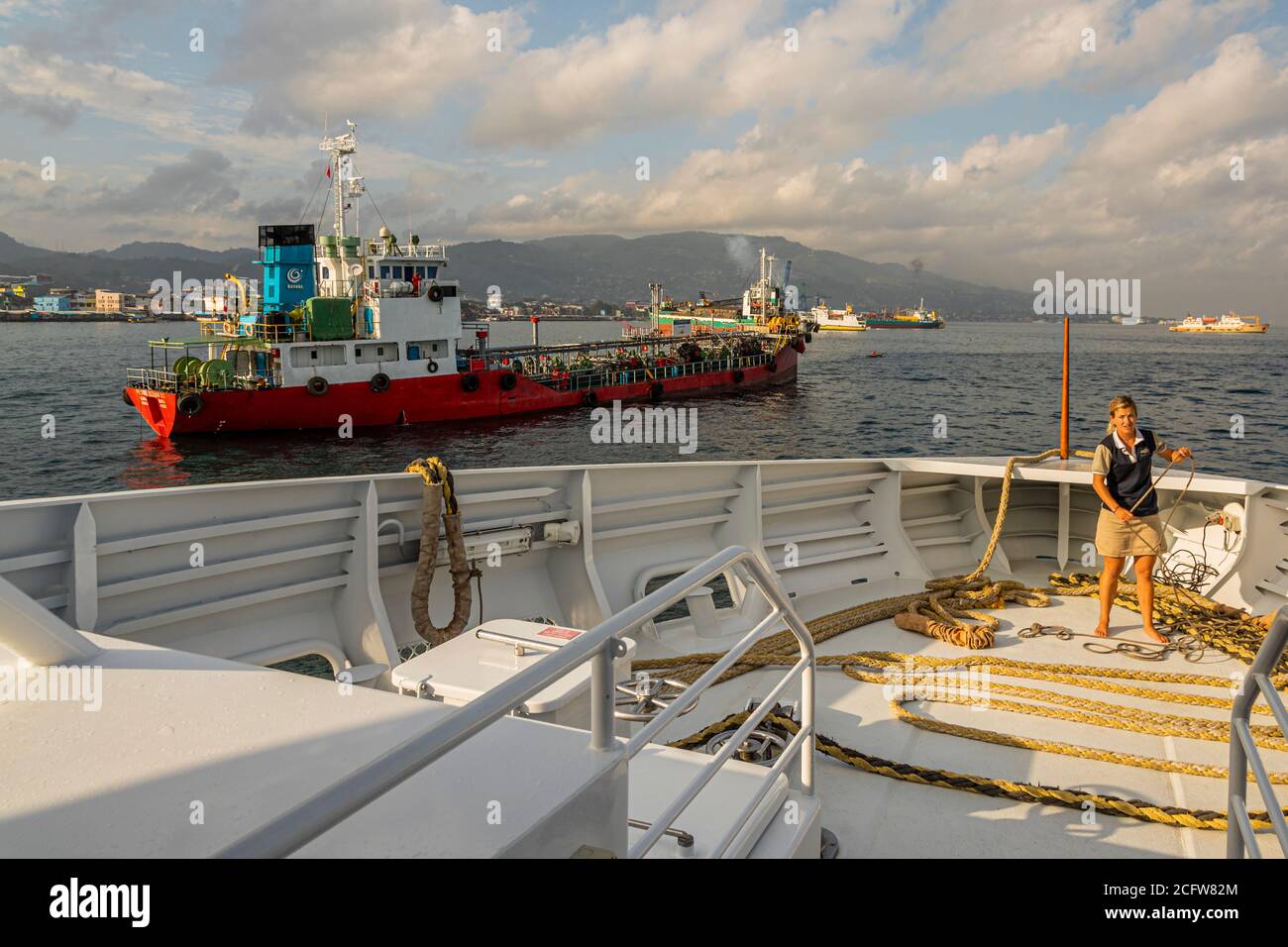 Un cisterna rossa giace al largo della città collinare di Ambon, Indonesia, mentre un membro dell'equipaggio avvolge le cime di ormeggio sul ponte di prua del True North Foto Stock