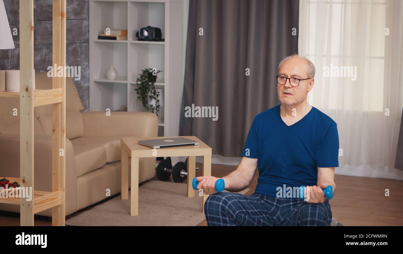 Pensionato che fa addestramento del bicipite sulla sfera di equilibrio in soggiorno. Anziano pensionato sano addestrando sport di salute a casa, esercitandosi attività di idoneità all'età anziana Foto Stock