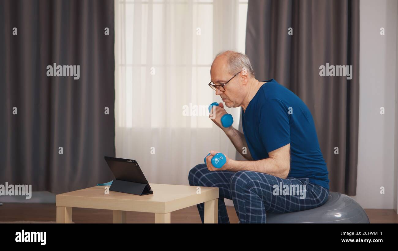 Anziani maschi anziani che si allenano con manubri seduti sulla palla svizzera durante l'allenamento online. Anziano pensionato sano addestrando sport di salute a casa, esercitandosi attività di idoneità all'età anziana Foto Stock