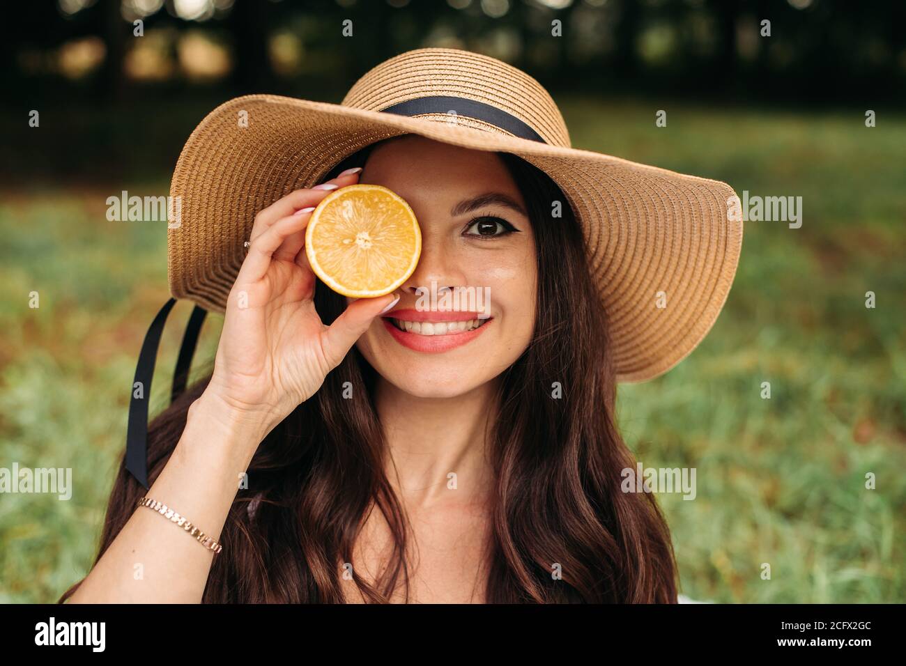 Foto ritratto di stock di bella ragazza con lunghi capelli marroni in elegante cappello estivo che tiene mezzo arancione in mano nel parco. Sorridendo alla macchina fotografica con happine Foto Stock