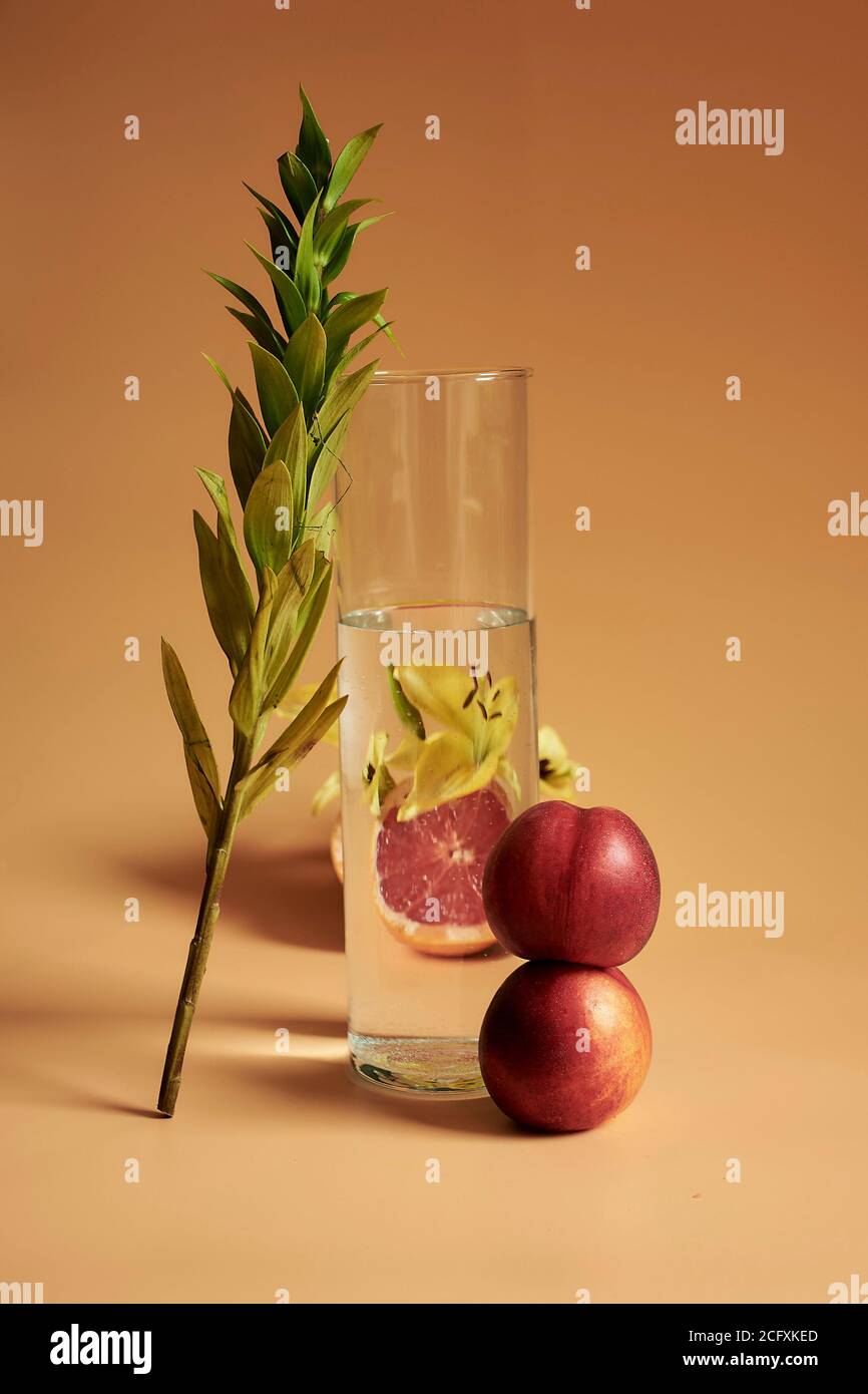 Una vita still di foglia che conduce su un bicchiere d'acqua con un fiore giallo e pesche su uno sfondo arancione che fa un look monocromatico. Foto Stock