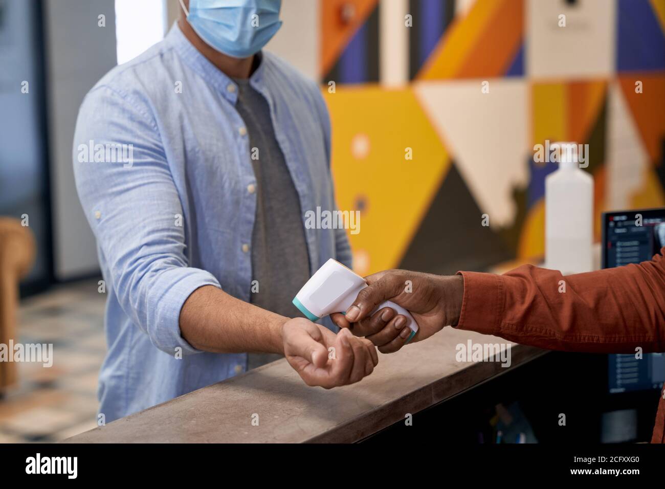 Controllo della temperatura sul lavoro, receptionist maschile africano indossando maschera medica utilizzando un termometro a infrarossi per misurare la temperatura corporea dell'ufficio Foto Stock