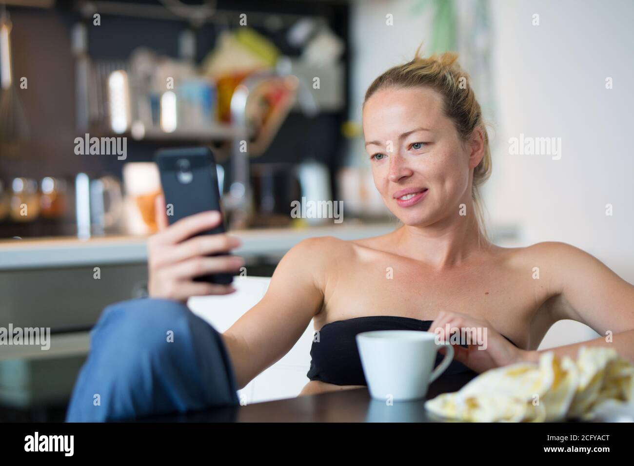 Giovane sorridente allegra donna soddisfatta all'interno della cucina domestica usando social media sul telefono cellulare per chattare e rimanere connessi con i suoi cari Foto Stock