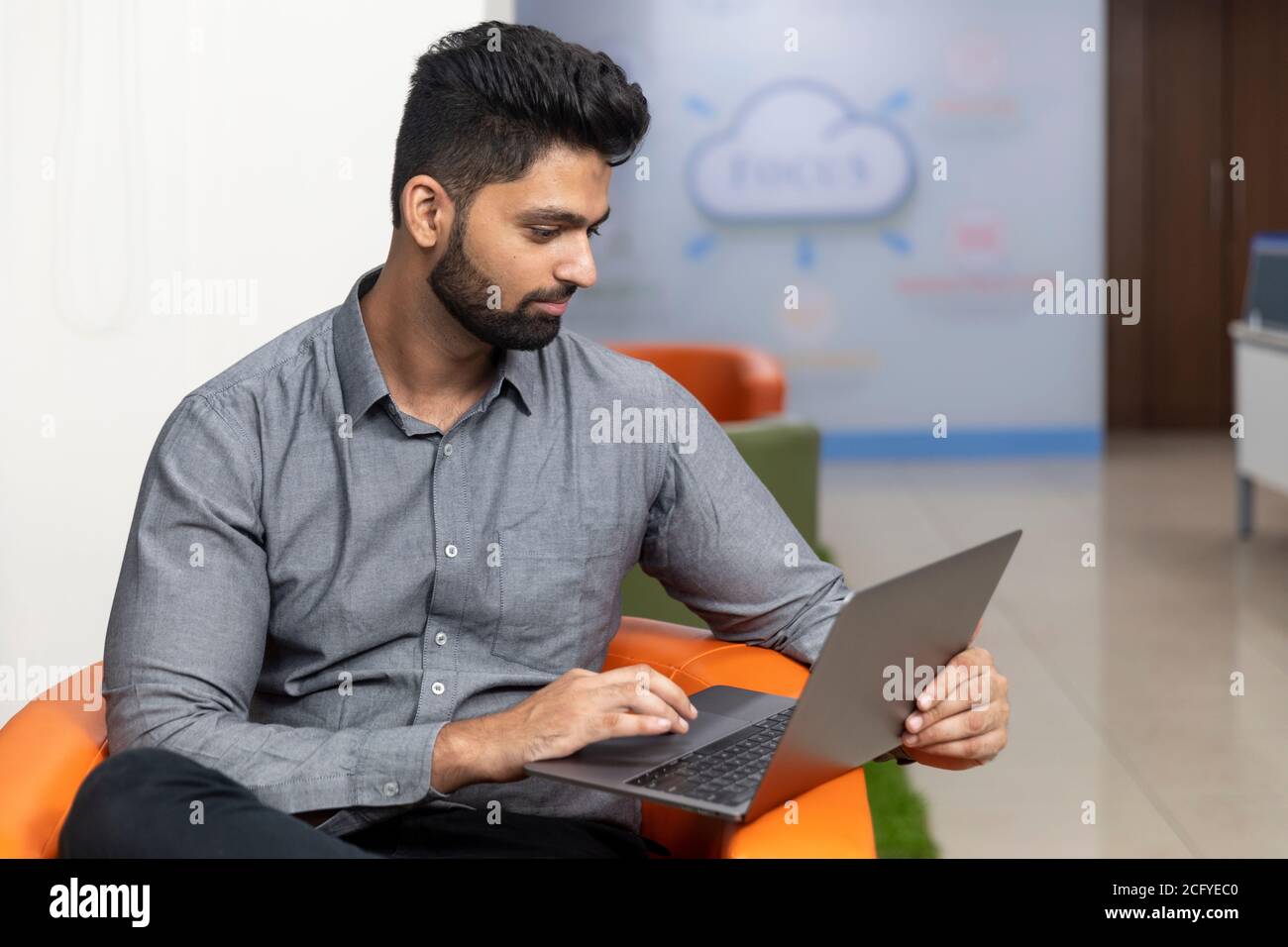 Ritratto di giovane uomo d'affari indiano di successo seduto sul divano e lavorando su un laptop, ambiente aziendale. Foto Stock