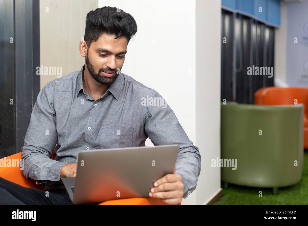 Ritratto di giovane uomo d'affari indiano di successo seduto sul divano e lavorando su un laptop, ambiente aziendale. Foto Stock
