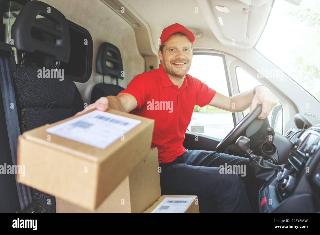 giovane sorridente addetto al servizio di consegna seduto in furgone e consegna scatole Foto Stock