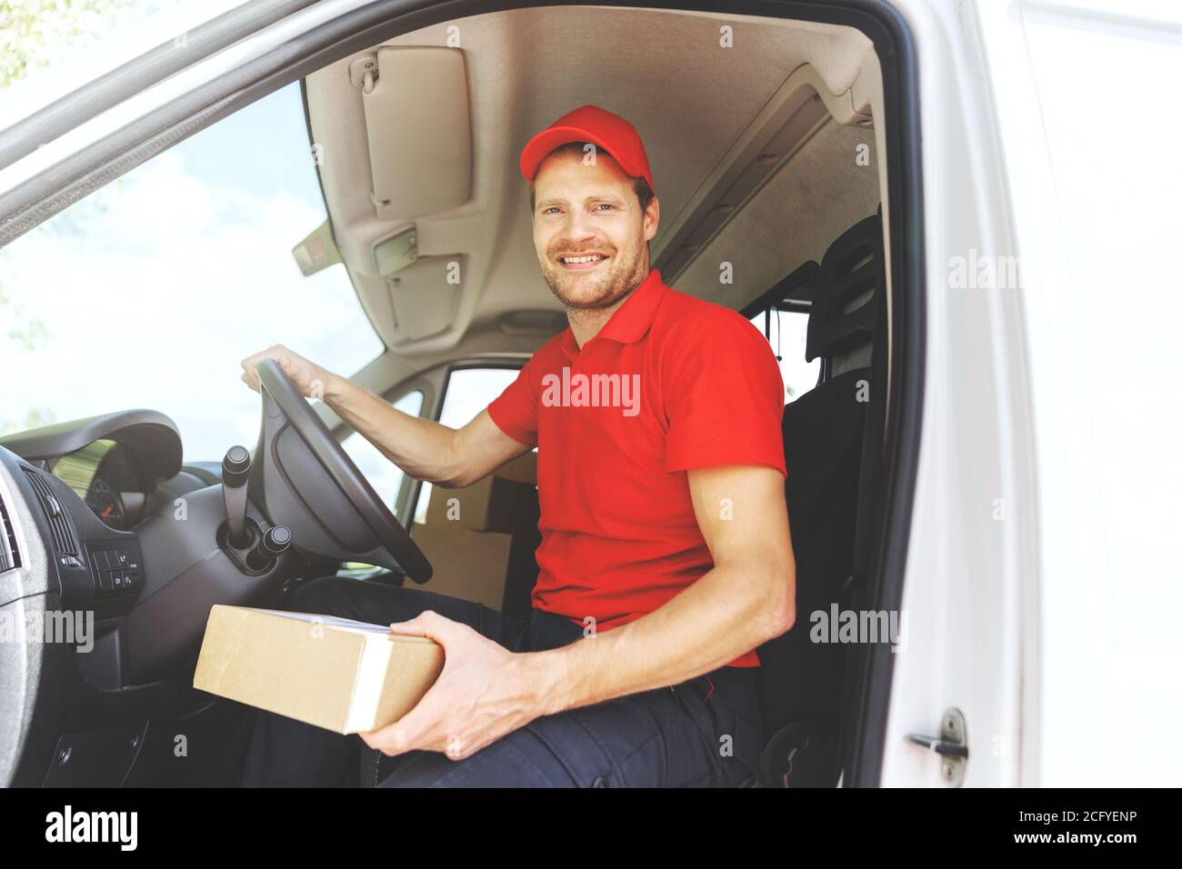 sorridente autista del servizio di trasporto in uniforme rossa seduto in pulmino con scatola in mano Foto Stock