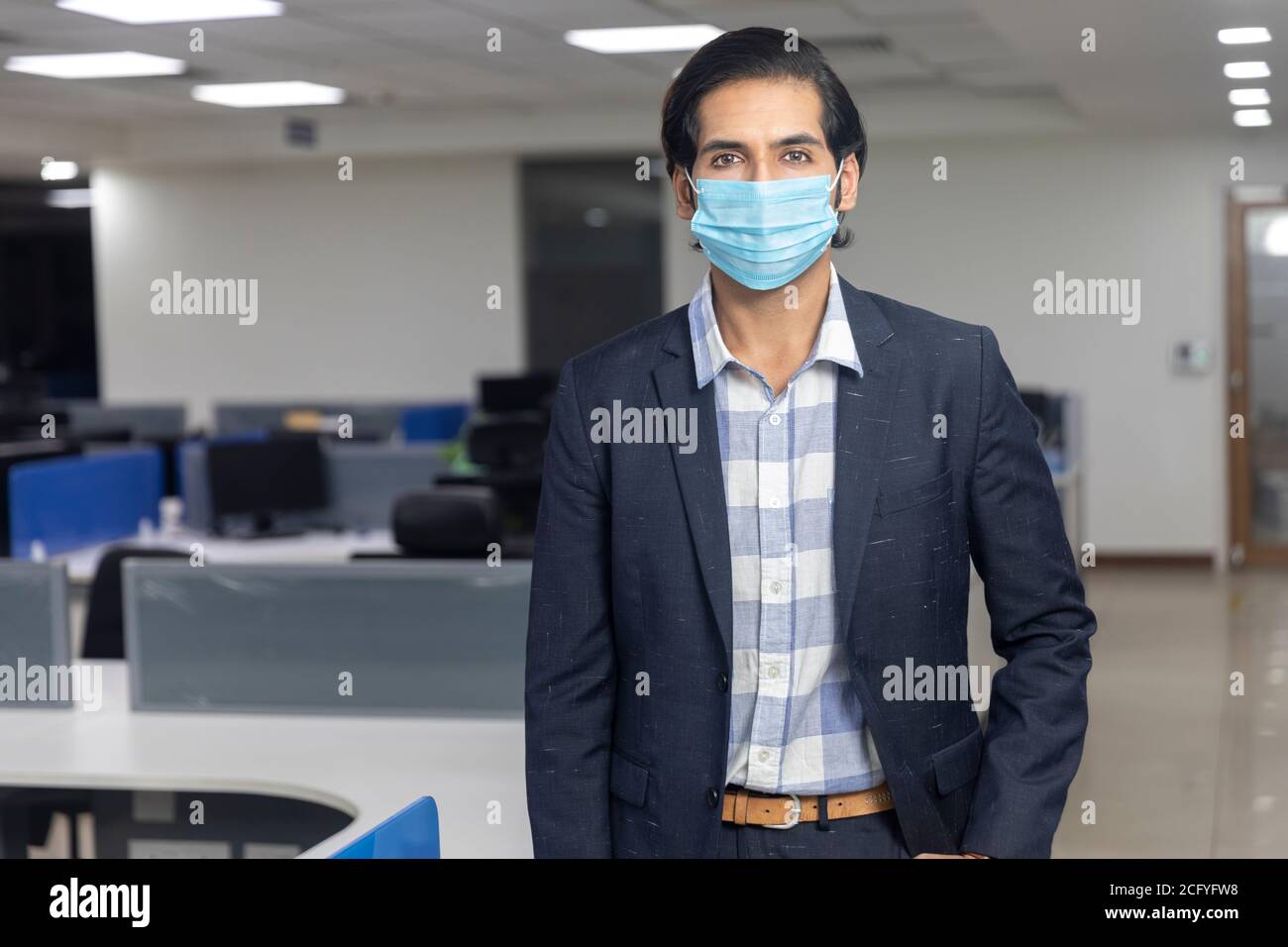 Ritratto di bel giovane uomo d'affari indiano che indossa maschera di protezione Covid, in piedi contro lo sfondo dell'ufficio. Ambiente aziendale. Foto Stock