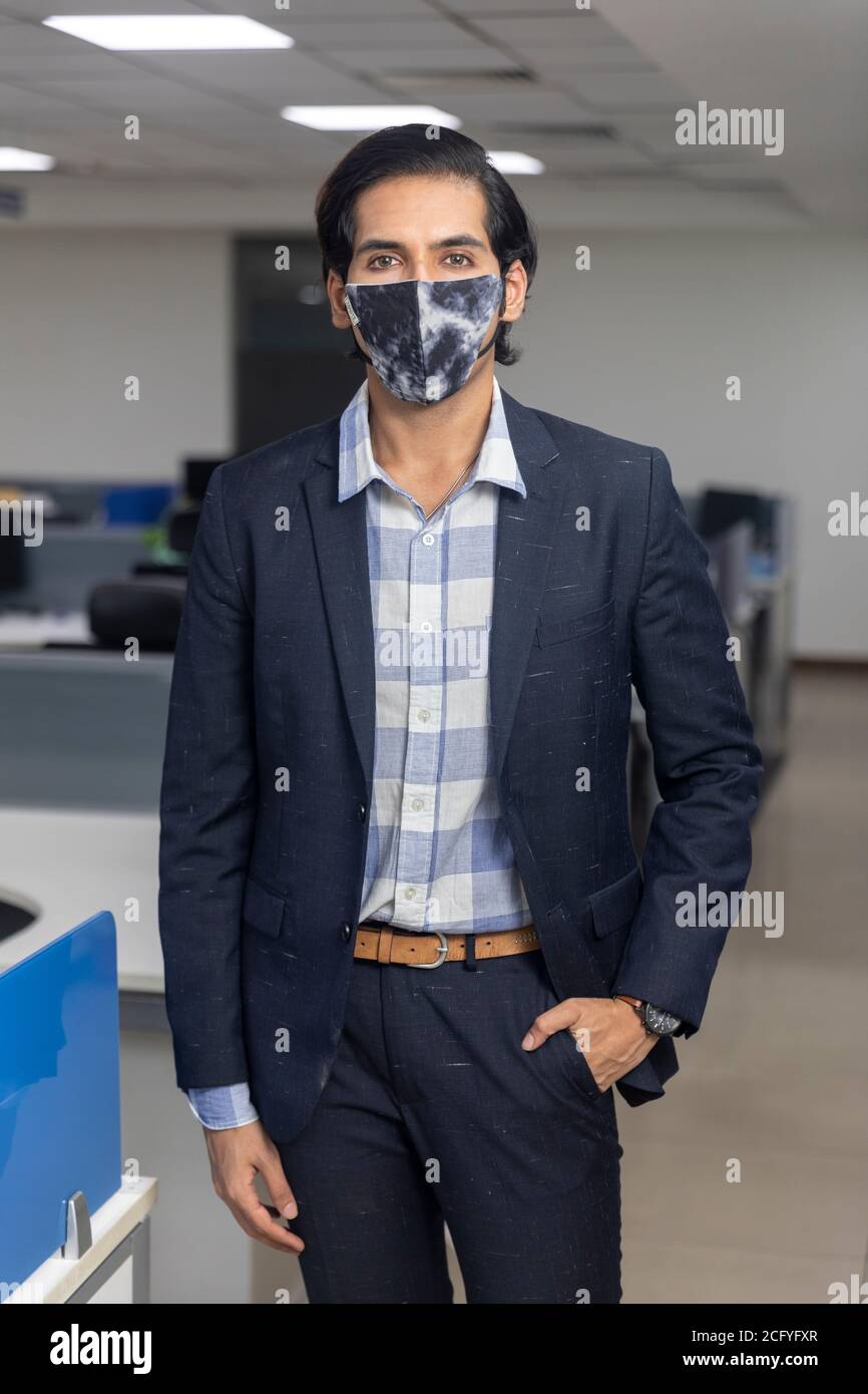 Ritratto di bel giovane uomo d'affari indiano che indossa maschera di protezione Covid, in piedi contro lo sfondo dell'ufficio. Ambiente aziendale. Foto Stock