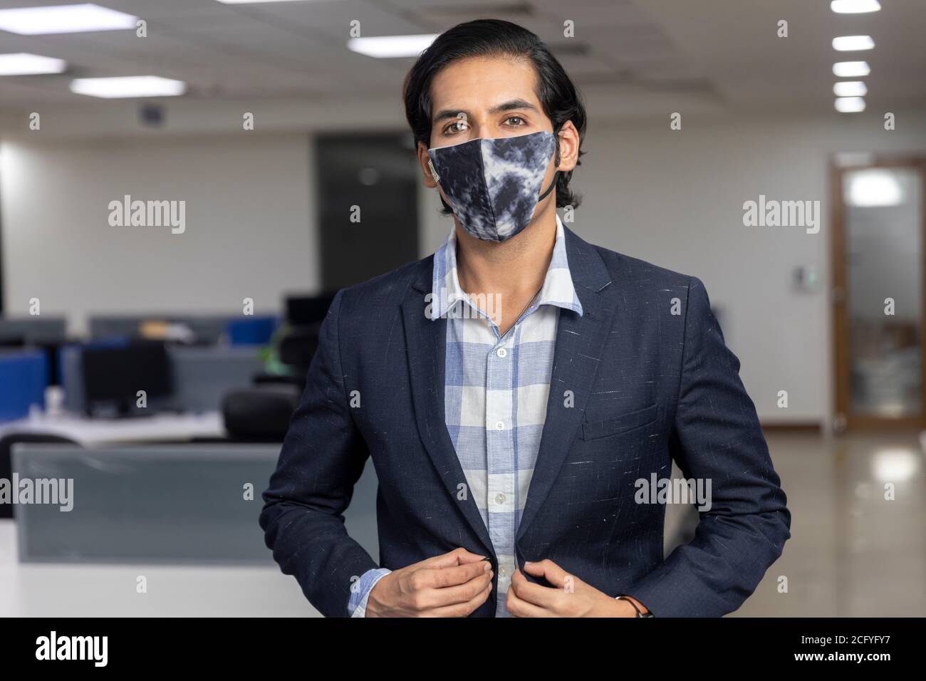 Ritratto di bel giovane uomo d'affari indiano che indossa maschera di protezione Covid, in piedi contro lo sfondo dell'ufficio. Ambiente aziendale. Foto Stock