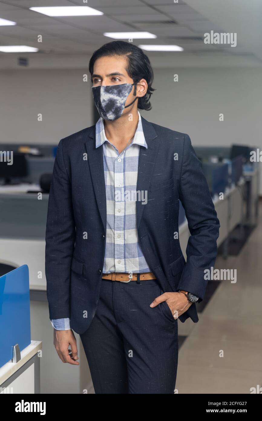 Ritratto di bel giovane uomo d'affari indiano che indossa maschera di protezione Covid, guardando lontano dalla fotocamera, ambiente aziendale ufficio. Foto Stock