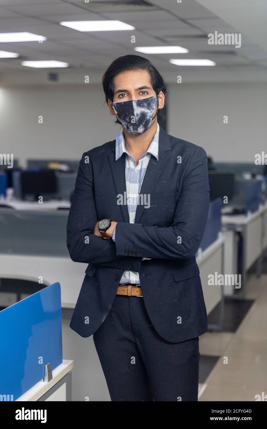 Ritratto di bel giovane uomo d'affari indiano che indossa maschera di protezione Covid, in piedi contro lo sfondo dell'ufficio. Ambiente aziendale. Foto Stock