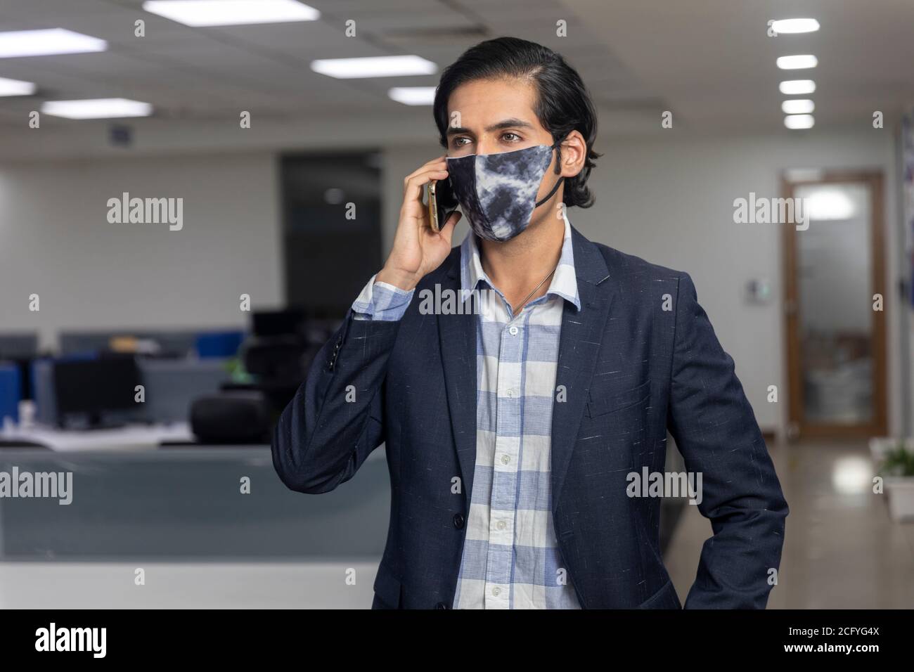 Ritratto di bel giovane uomo d'affari indiano che indossa una maschera di protezione Covid, parlando sul suo telefono cellulare, ambiente aziendale. Foto Stock