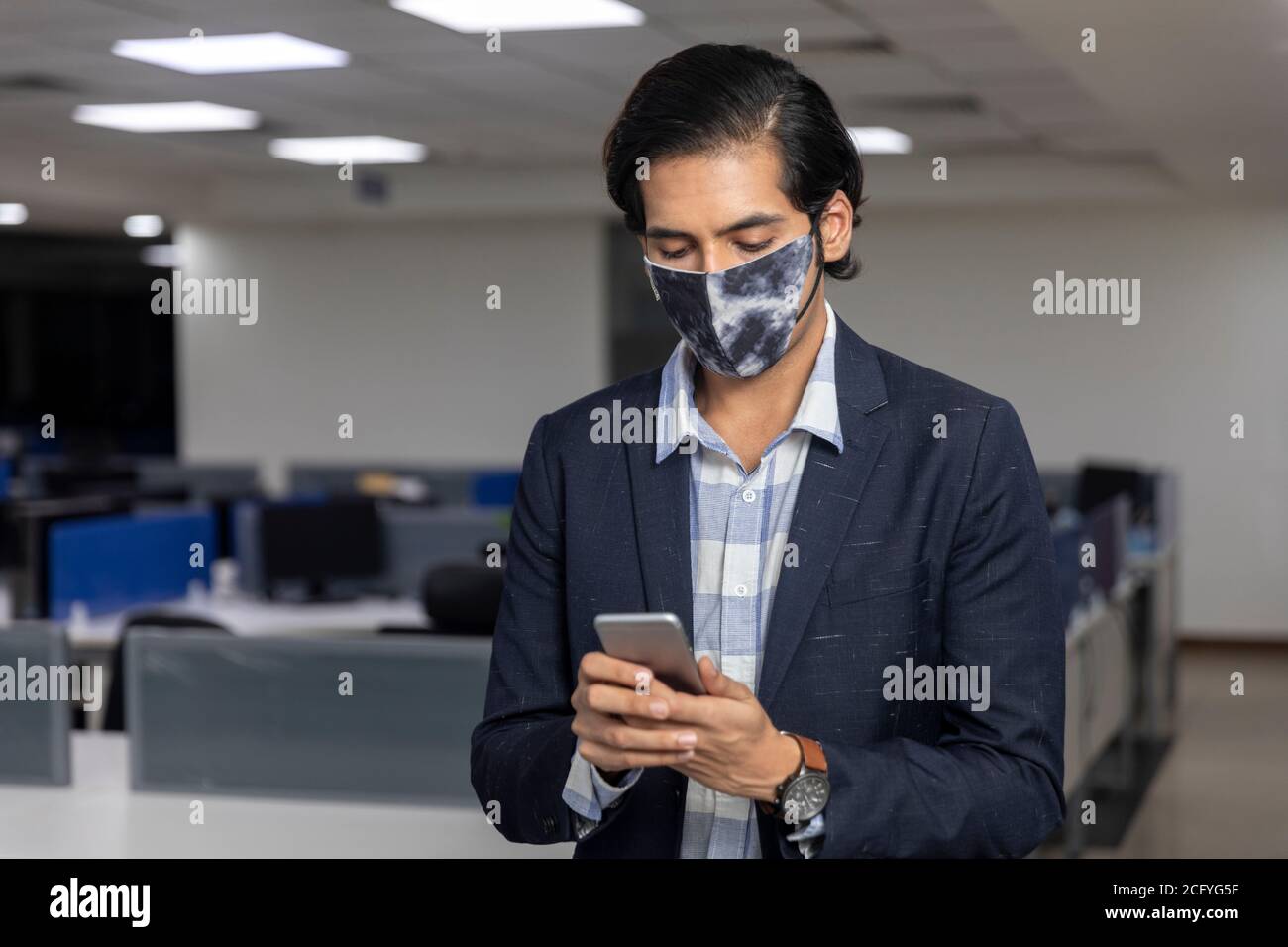 Ritratto di bel giovane uomo d'affari indiano indossare maschera di protezione Covid e controllare il suo telefono cellulare. Ambiente aziendale. Foto Stock