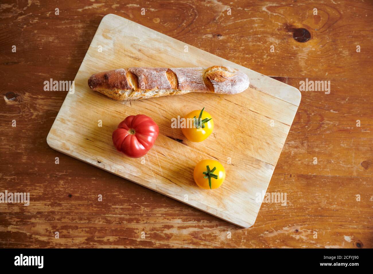 Auf einem hellen Holzschneidebrett liegen ein Baguette, 2 gelbe Tomaten und eine rote Tomaten die selbst auf dem Balkon gezogen wurden. Foto Stock