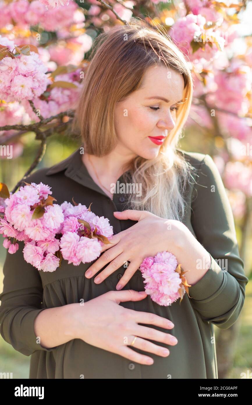 Donna incinta bionda incinta in primavera nel parco con alberi sakura, profondità bassa di campo di gravidanza bagliore Foto Stock