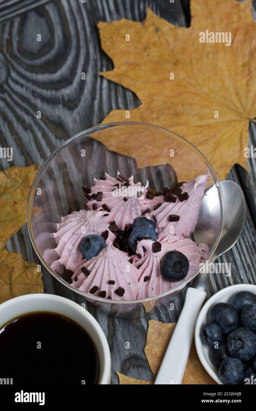 Zephyr in un bicchiere. Con biscotto e mirtilli. Accanto si trova una tazza di caffè e un piatto di mirtilli. Tra le foglie d'acero. Su tavole di pino pai Foto Stock