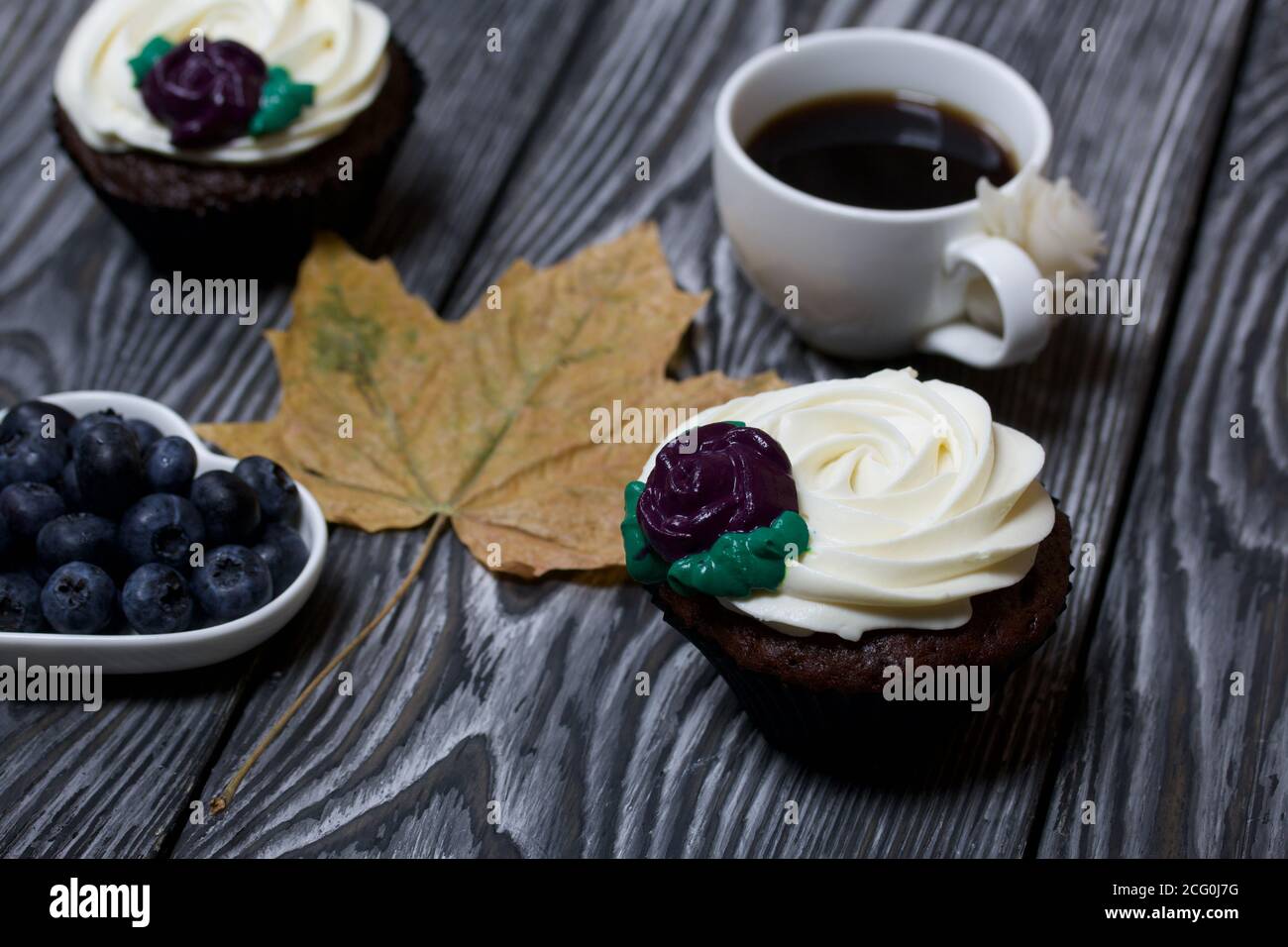Muffin al cioccolato con fiore alla crema. Una tazza di caffè nero. Piatto con mirtilli. Foglia di acero essiccata. Si trova su tavole di pino dipinte in bianco e nero. Foto Stock